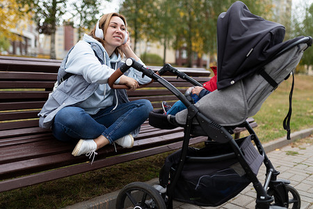 走路听音乐摄影照片_一位年轻的母亲推着婴儿车走路时坐在长椅上，戴着耳机听音乐