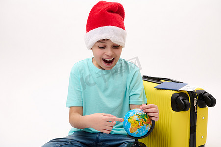 坐在黄色手提箱旁边的戴着圣诞帽的顽皮少年指着地球上的一个旅游目的地。