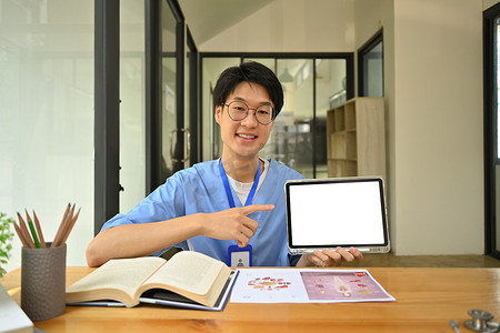 年轻的亚洲男医学生身穿蓝色磨砂膏，展示数字平板电脑并指着空白屏幕