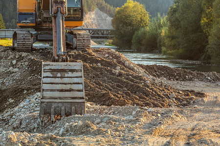 小黄色挖掘机在河边的一堆岩石和石头上，地面上挖掘机铲斗的细节。