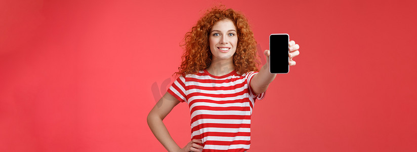 自信漂亮的红发卷发女性展示酷炫的应用程序手持智能手机扩展手显手机屏幕微笑自信推荐订阅她的博客社交媒体