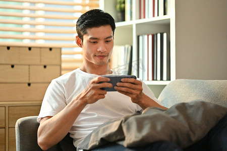 工作玩手机摄影照片_快乐的千禧一代男人躺在沙发上玩智能手机游戏。技术、娱乐和休闲活动