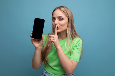 蓝色背景竖摄影照片_穿着休闲装的金发美女竖着拿着智能手机，蓝色背景上用手指向前遮住嘴巴的屏幕模型