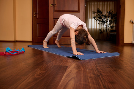 狗拉伸摄影照片_可爱的运动型小女孩以下犬式伸展身体，在舒适的室内练习瑜伽