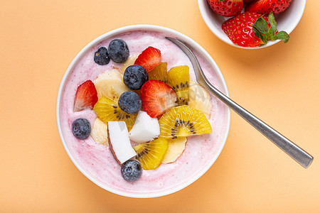 健康早餐或甜点酸奶碗，配以新鲜香蕉、草莓、蓝莓、可可、猕猴桃，以最小的柔和纸背景和勺子为顶视图