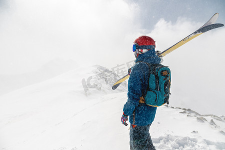 降雪期间，在山的背景下，男人背对着镜头站着，肩上扛着高山滑雪板