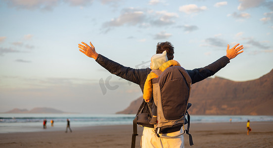 婴儿背包摄影照片_日落时，年轻的父亲在西班牙兰萨罗特岛 Famara 多风的沙滩上，一边享受纯净的大自然，一边背着他的婴儿男婴儿子背着背包，举起双手向天空。