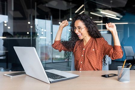 西班牙裔女商人庆祝胜利成功，办公室里卷发的员工阅读好消息，在办公室工作时使用笔记本电脑举起手和快乐的胜利手势
