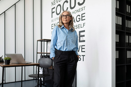 身穿蓝色衬衫的女高管站在办公室中央，望向远方，白人中年老年女商人在工作概念