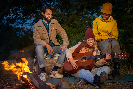 三个快乐的不同朋友玩音乐，晚上在大自然中享受篝火露营