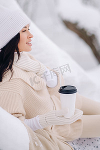 一个穿着米色开衫和白帽子的漂亮女孩在湖边的雪冬森林里喝茶