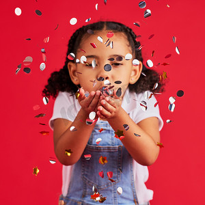 彩活动摄影照片_年轻的女孩，孩子们在聚会和庆祝活动中吹彩纸屑，银色和闪光在红色背景下被隔离。