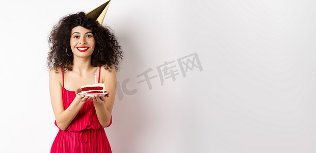 生日许愿摄影照片_穿着红色连衣裙、戴着派对帽庆祝生日、拿着生日蛋糕许愿、对着镜头微笑、站在白色背景上的漂亮女人