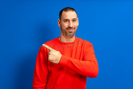 路径指示摄影照片_留着胡须的西班牙裔男子身穿红色毛衣，对着镜头微笑，同时指着蓝色背景中孤立的一侧，指示要遵循的路径或方向。