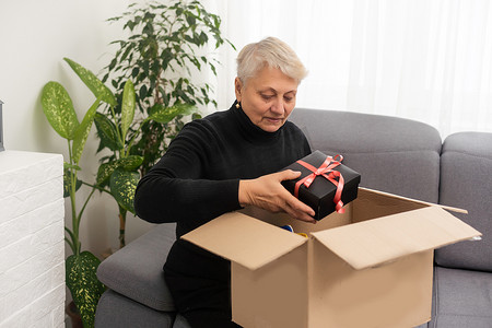 微笑的老年女性顾客在家里收到邮寄包裹，快乐的老奶奶拿着打开的纸箱坐在客厅的沙发上，网上购物订单快速快递概念。