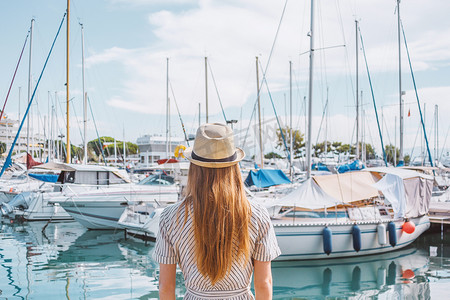 小船背景摄影照片_戴着帽子的金发女人背对着大海、游艇和小船背景站着。