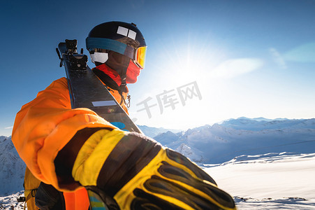 扛在肩上摄影照片_在阳光明媚的天空背景下，冬季山区的滑雪者，从下方广角，一名男子将滑雪板扛在肩上