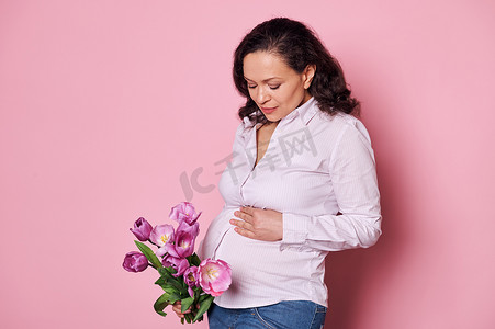 高兴的孕妇抚摸着她的肚子，第一次感觉到婴儿在踢，拿着一束郁金香，粉红色的背景。