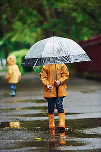 两个带雨伞的孩子穿着黄色防水斗篷和靴子在雨后一起在户外玩耍