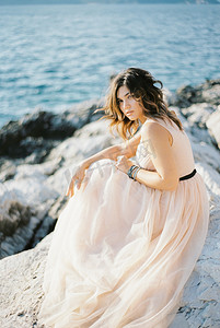 头发凌乱摄影照片_穿着米色连衣裙、头发凌乱的新娘坐在海边的岩石上
