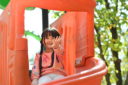 阳光明媚的早晨，在被绿树环绕的公园操场上玩乐的快乐亚洲小女孩