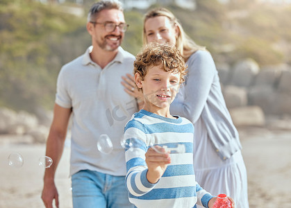 快乐的白人小男孩一边吹、捉和弹出肥皂泡，一边与父母一起在海滩度过轻松有趣的家庭暑假，享受美好时光。