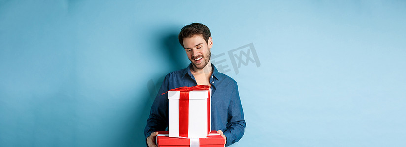 情人节浪漫蓝色摄影照片_留着胡子的浪漫年轻人，在情人节看着礼盒很开心，给情人送礼物，站在蓝色背景上