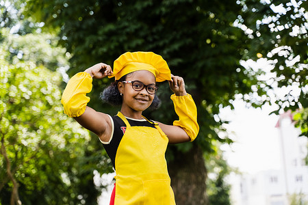 身穿厨师帽和黄色围裙制服的非洲黑人儿童厨师女孩摸着她的厨师帽。