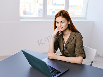 一位女士坐在明亮办公室的桌子旁，窗户背景是一台笔记本电脑