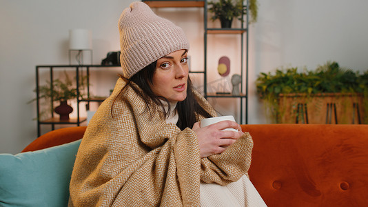 生病的不健康女人戴着裹着格子花呢的帽子，坐在沙发上喝着热茶冻得发抖
