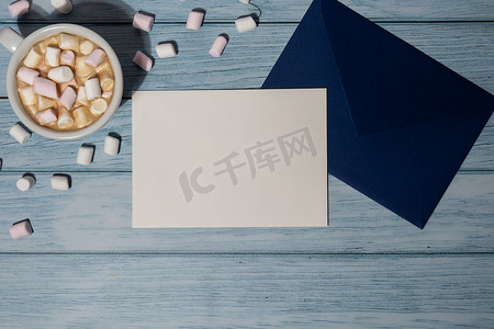 空的问候语或邀请卡模拟了蓝色信封，白色的咖啡和棉花糖在木制的蓝色背景上。