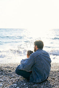 穿着牛仔夹克的爸爸坐在卵石滩上，膝上抱着一个小女孩。