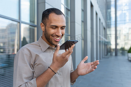 微笑的西班牙裔商人录制语音信息，办公楼外快乐的人在智能手机上阅读指令进行人工智能搜索