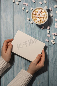 女手拿着空的问候语或邀请卡，在木制蓝色背景上用白杯咖啡和棉花糖进行模拟。