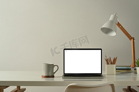 在最小的家庭办公室中，笔记本电脑、咖啡杯、铅笔架和白桌上的灯的正面视图