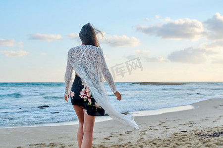 飘动摄影照片_穿着时尚浪漫服装的年轻女性沿着海滩散步的背影