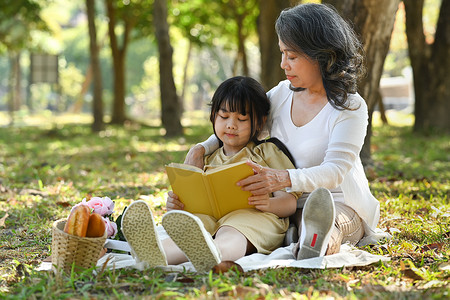 慈爱的祖母拥抱学龄前小孙女，在阳光明媚的夏日花园里阅读童话故事