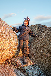 傍晚的阳光下，一个戴着针织帽的微笑可爱男孩站在金色的稻草堆上。