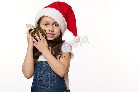一个戴着圣诞老人帽子的可爱小女孩的白色肖像，轻轻地拥抱着她的圣诞礼物，看着相机。
