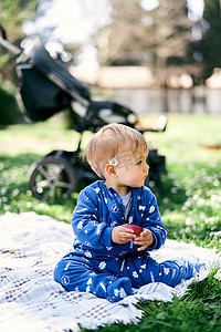 身穿蓝色工装裤、耳后有一朵雏菊的小孩坐在草坪上的毯子上，手里拿着一个苹果，背景是婴儿车，看着旁边