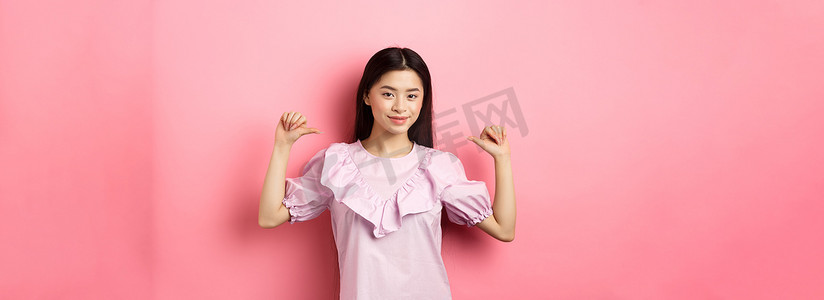 自信的亚洲女孩微笑着指着自己，自我提升个人成就，穿着粉色背景的裙子
