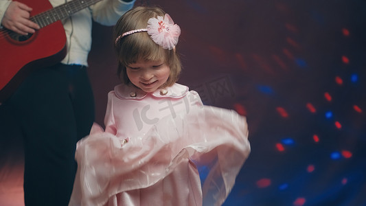穿着复古连衣裙的小女孩在舞台上跳舞，她的父亲弹着原声吉他