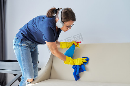 家庭清洁，戴耳机的女人用抹布和用洗涤剂洗皮沙发的喷雾