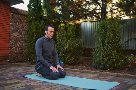 运动健身垫摄影照片_中年活跃男子瑜伽士坐在健身垫上，在户外练习呼吸和放松运动。