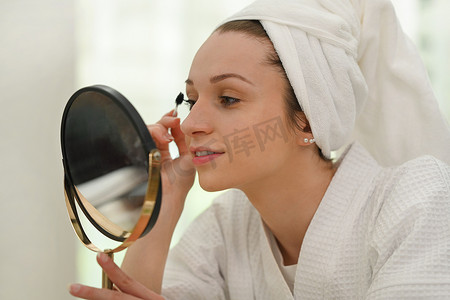 美丽的白种女人穿着浴袍在睫毛上涂睫毛膏的照片。