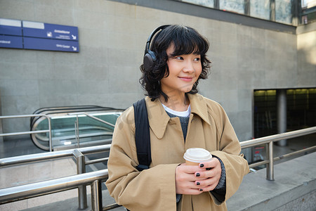 年轻学生的画像，戴耳机的女孩，喝咖啡，背着背包站在街上，上大学或学院，开心地微笑