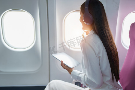 年轻的亚洲美女乘客坐在商务舱豪华飞机上，同时戴着无线耳机在飞行期间放松和使用平板电脑