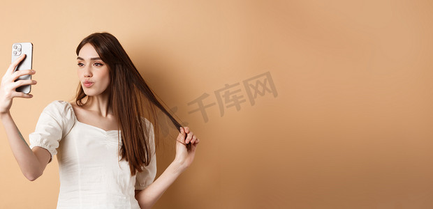 时尚的年轻女性玩头发，在智能手机上自拍，为社交媒体拍照，站在米色背景中