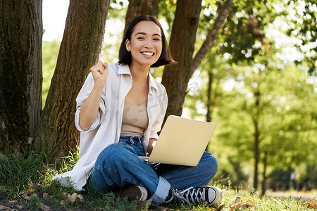 在阳光明媚的绿色公园里，热情的亚洲年轻女孩拿着笔记本电脑坐在树旁，庆祝、胜利和微笑