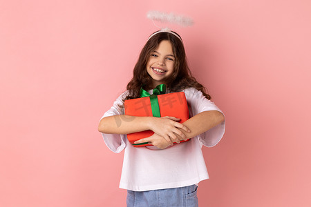 微笑天使摄影照片_天使光环中的小女孩拥抱包裹着的盒子，享受生日礼物，对礼物感到满意。